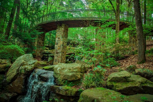 A stone bridge arches over a small waterfall, surrounded by lush green trees and rocky terrain.