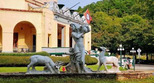 A statue of a woman holding a fish, flanked by two deer, in front of a historic building surrounded by greenery.