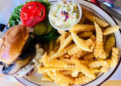 A plate with a burger topped with cheese and mushrooms, lettuce, tomato, pickles, coleslaw, and crinkle-cut fries.