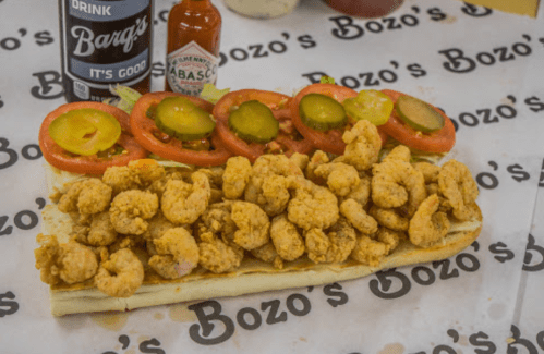 A large sandwich filled with fried shrimp, lettuce, tomatoes, pickles, and bottles of Barq's soda and Tabasco sauce.