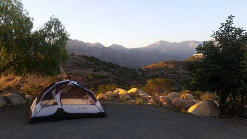 A tent set up on a gravel area with mountains and trees in the background under a clear sky.