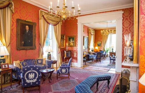 Elegant room with red wallpaper, ornate chandeliers, classic furniture, and framed portraits. Cozy and inviting atmosphere.