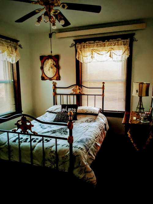 Cozy bedroom with a metal bed, decorative pillows, vintage lamp, and two windows with blinds. Warm, inviting atmosphere.