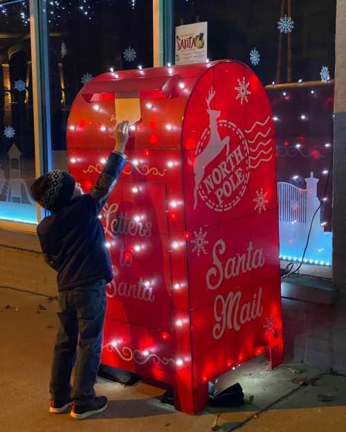 A child in a winter hat places a letter into a brightly lit red mailbox labeled "Santa Mail" at night.