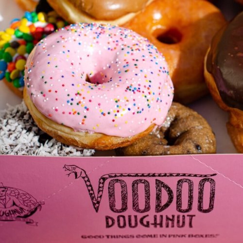 A colorful assortment of doughnuts, including a pink frosted one with sprinkles, displayed with a Voodoo Doughnut box.
