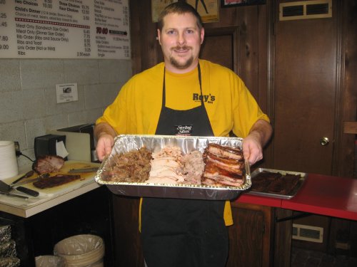 A smiling man in a yellow shirt holds a tray of assorted barbecue meats in a restaurant setting.
