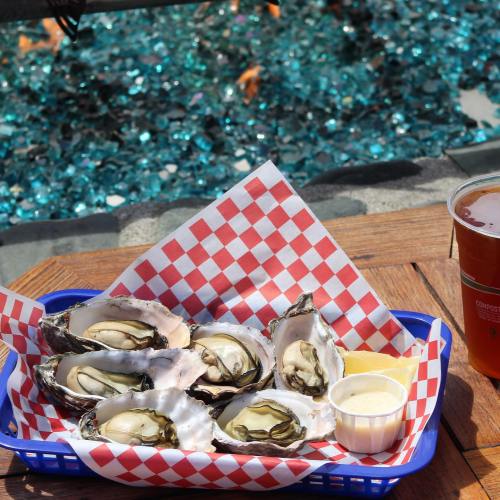 A basket of fresh oysters on a checkered paper, served with lemon and dipping sauce, alongside a glass of beer.