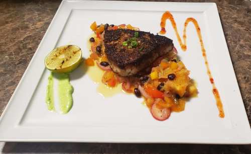 A beautifully plated blackened fish fillet with mango salsa, lime, and colorful sauces on a white square plate.