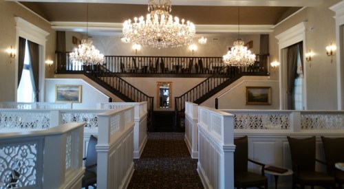 Elegant interior of a staircase with chandeliers, framed artwork, and decorative railings in a well-lit space.