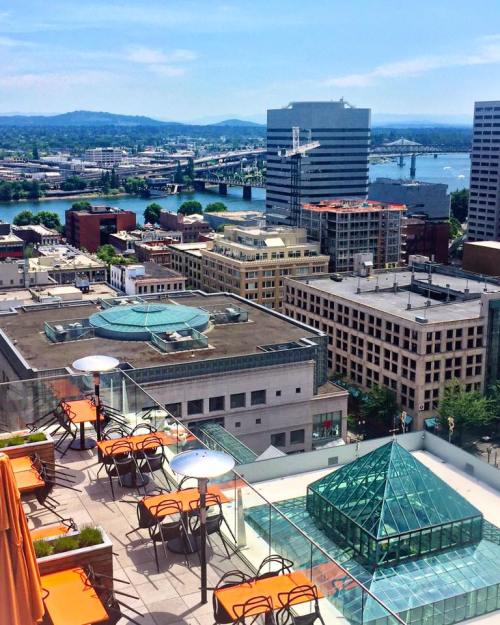 Aerial view of a cityscape featuring buildings, a river, and mountains in the background under a clear blue sky.
