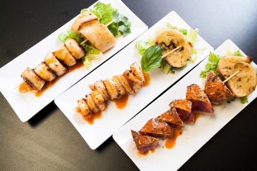 Three plates of grilled skewers featuring shrimp, chicken, and vegetables, garnished with lettuce and drizzled with sauce.