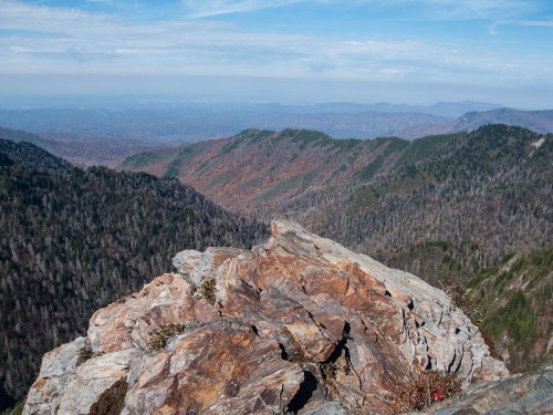 A rocky outcrop overlooks a vast valley filled with autumn-colored trees and distant mountains under a blue sky.