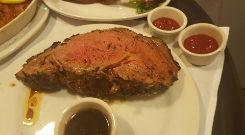 A large, juicy slice of prime rib on a plate, accompanied by two small bowls of sauce.