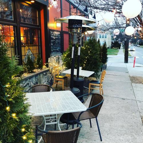Outdoor dining area with tables, chairs, and a heater, decorated with string lights and lanterns along a sidewalk.