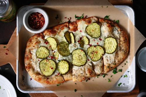 A flatbread topped with cheese, pickles, and herbs, served on parchment paper with a side of red pepper flakes.