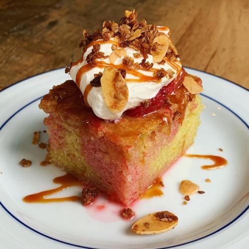 A slice of colorful cake topped with whipped cream, caramel drizzle, granola, and sliced almonds on a white plate.