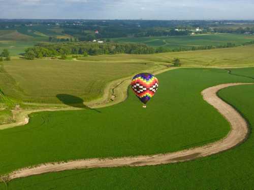 A colorful hot air balloon floats over lush green fields and winding paths under a clear blue sky.