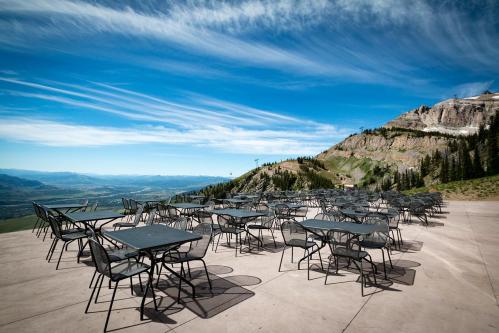 Empty outdoor dining area with metal tables and chairs, set against a backdrop of mountains and a blue sky.