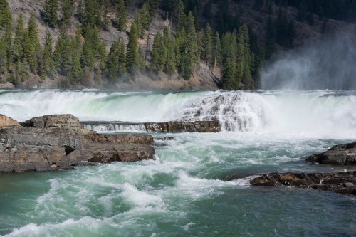 A powerful waterfall cascades over rocky terrain, surrounded by lush green trees and mist rising from the water.