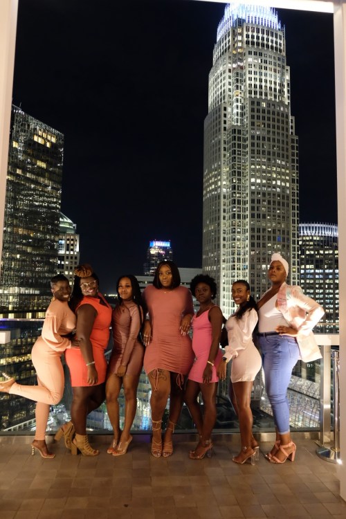 A group of seven women in stylish outfits pose together at night with a city skyline in the background.