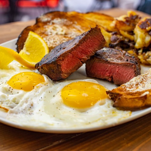 A plate of breakfast featuring two sunny-side-up eggs, steak slices, toast, and lemon wedges.