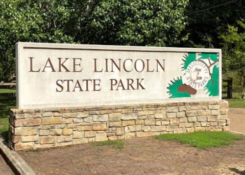 Sign for Lake Lincoln State Park, featuring stone base and green decorative elements, surrounded by trees.