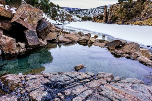 A serene natural hot spring surrounded by rocky terrain and snow-covered mountains in the background.