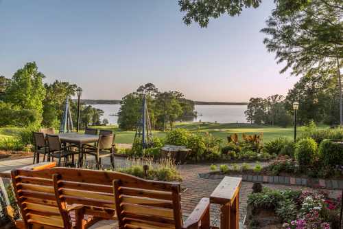 Scenic view of a lake from a landscaped patio with wooden seating and lush greenery under a clear sky.