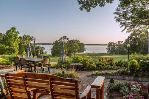 Scenic view of a lake from a landscaped patio with wooden seating and lush greenery under a clear sky.