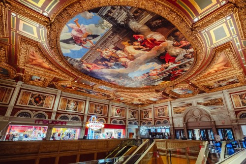 Ornate ceiling mural depicting classical scenes in a lavishly decorated indoor space, with shops visible below.