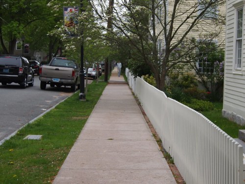 A sidewalk lined with a white picket fence, trees, and parked cars on a quiet street.