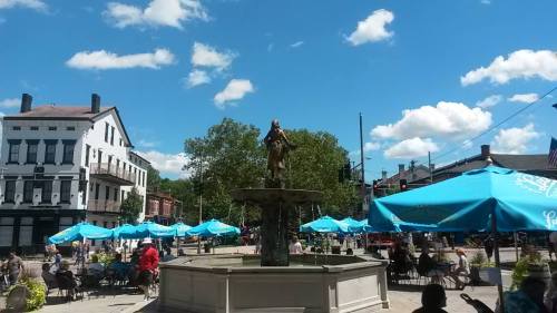A sunny plaza with a fountain and outdoor seating under blue umbrellas, surrounded by trees and historic buildings.