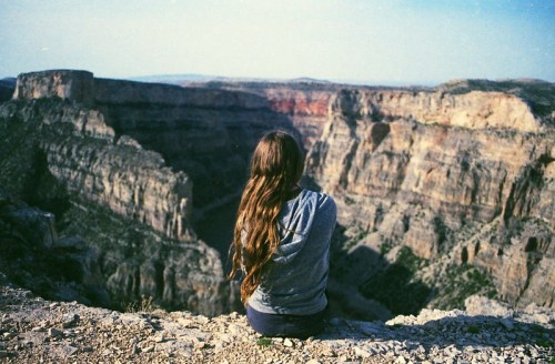 A person with long hair sits on a rocky ledge, gazing at a vast canyon landscape under a clear sky.