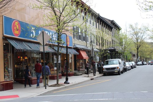 A street scene featuring shops, pedestrians, and parked cars, with trees lining the sidewalk on a cloudy day.