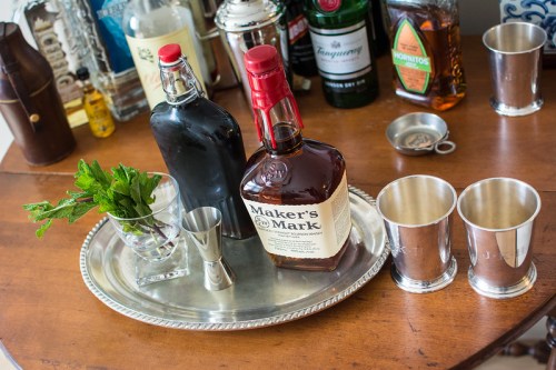 A silver tray holds a bottle of Maker's Mark bourbon, a black bottle, mint leaves, a jigger, and two silver cups.