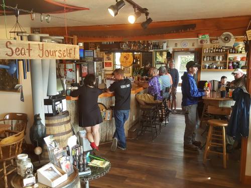 A cozy bar scene with patrons at the counter and tables, featuring a "Seat Yourself!" sign and a rustic decor.