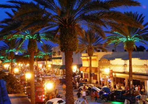 A vibrant street scene at night, lined with palm trees and illuminated shops, bustling with people and classic cars.