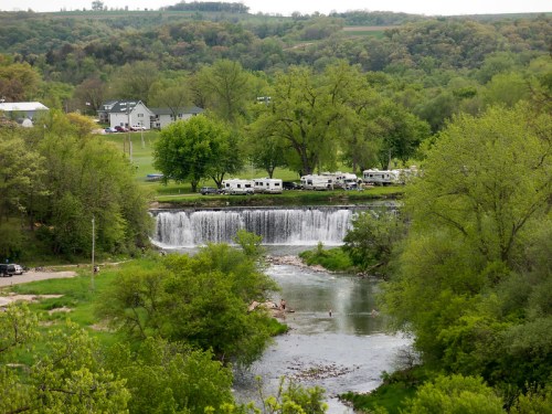 A scenic view of a waterfall surrounded by lush greenery and RVs parked nearby along a riverbank.