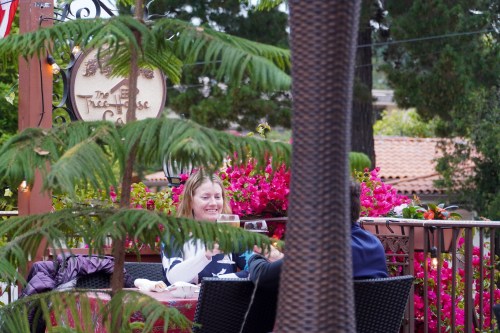 A woman smiles while sitting at a café table surrounded by vibrant flowers and greenery.