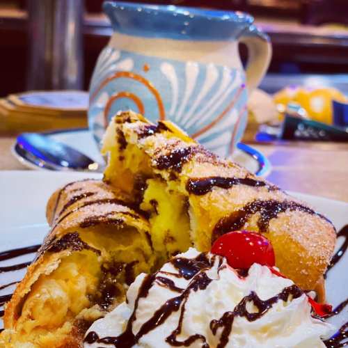 A plate of dessert featuring a chocolate-drizzled pastry, whipped cream, and cherries, with a decorative mug in the background.