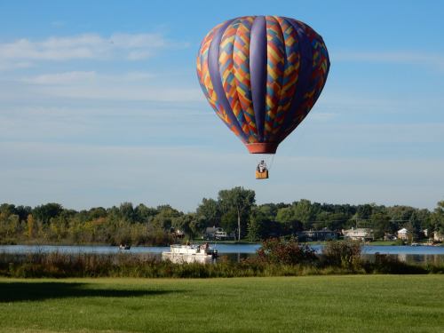 A colorful hot air balloon floats above a serene lake, with boats and lush greenery in the background.