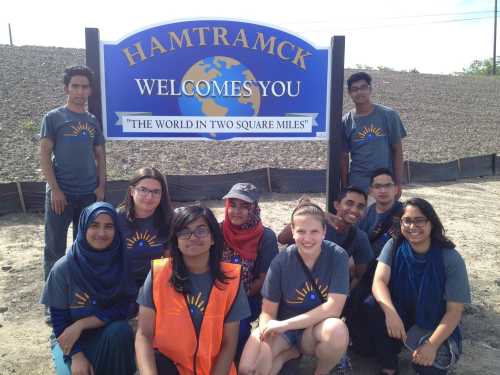 A diverse group of people poses in front of a "Hamtramck Welcomes You" sign, smiling and wearing matching shirts.