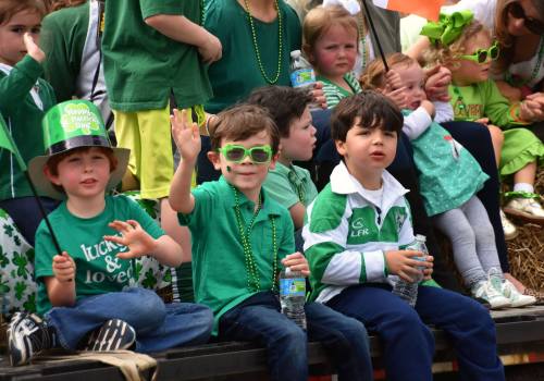Three children in green outfits celebrate St. Patrick's Day, waving and smiling while sitting on a float.