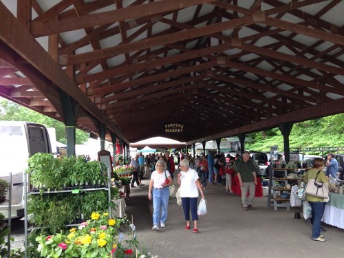 A bustling farmer's market under a wooden pavilion, with vendors and shoppers browsing fresh produce and flowers.