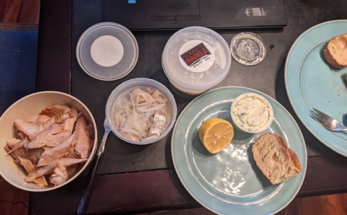 A table with plates of sliced fish, onions, lemon, cream cheese, and a piece of bread, alongside a laptop.