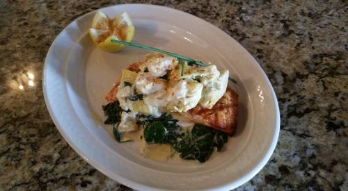 A plate of salmon topped with creamy artichokes and greens, served with a lemon wedge on a granite countertop.