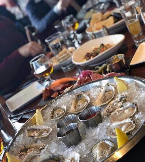 A platter of oysters on ice with lemon wedges, surrounded by various dishes and drinks on a dining table.