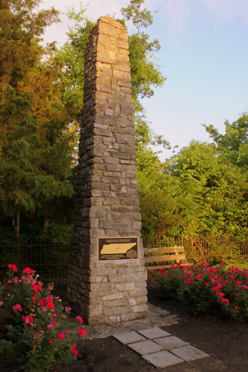 A tall stone monument surrounded by greenery and flowers, featuring a plaque at its base.