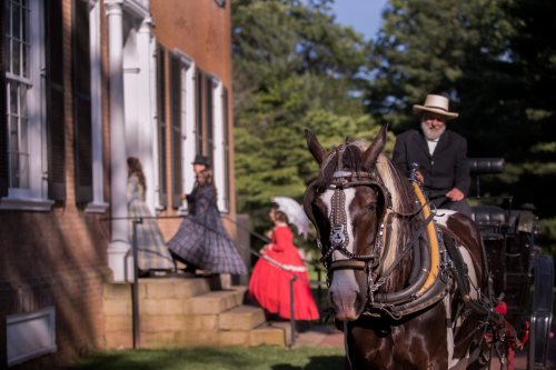 A man in a hat rides a horse near a historic building, while women in period dresses walk nearby.