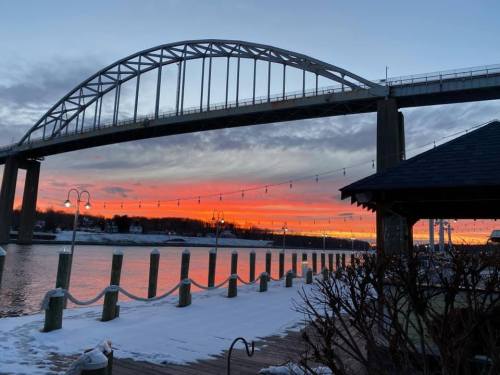 A bridge spans a river at sunset, with vibrant orange and purple skies reflecting on the water and snow-covered ground.
